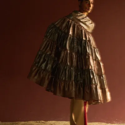 An avant-garde editorial look featuring a voluminous, tiered metallic cape. The styling creates a dramatic, sculptural silhouette, with a single red flower in the hair adding a point of sharp contrast against the moody, textured backdrop.