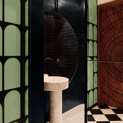 This powder room, attached to an entertainment den in Bangalore, has a bold, moody atmosphere. It features black tiles, checkered flooring, a carved wood door, and a sculptural pedestal marble sink.