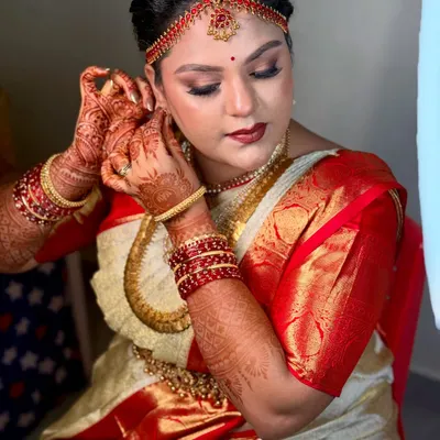 A candid moment of a bride getting ready, putting on her earrings. The makeup is set for a classic South Indian muhurtham look, with defined eyes and a bold lip to match her red and cream silk saree.