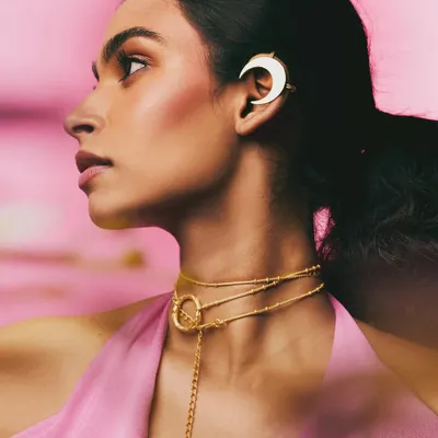 A profile shot for the Batoki Art jewelry campaign, featuring a delicate moon-shaped ear cuff and layered gold necklace. The soft pink background and gentle lighting create a romantic, feminine mood.
