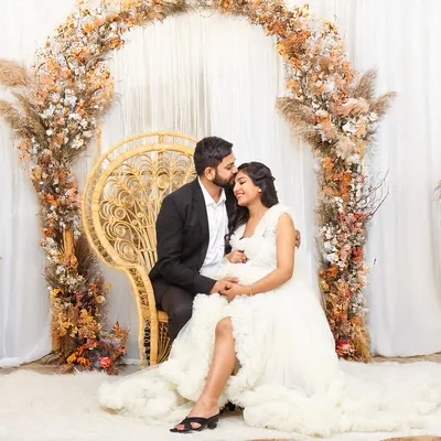 An expecting couple seated in a grand peacock chair, surrounded by a beautiful arch of pampas grass and autumn florals. This dreamy studio setup creates a warm and romantic atmosphere.