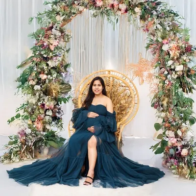 A stunning solo portrait of a mother-to-be seated on a wicker peacock chair, framed by a beautiful floral arch. The rich blue of her gown and the lush flowers create an enchanting and elegant scene.