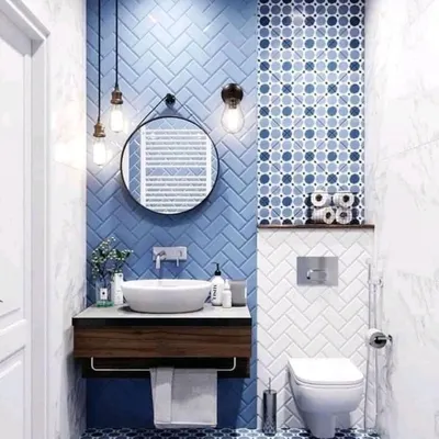 A vibrant and playful bathroom design featuring blue subway tiles in a herringbone pattern and complementary patterned floor tiles. The round mirror and hanging Edison bulbs add a touch of industrial-chic style.