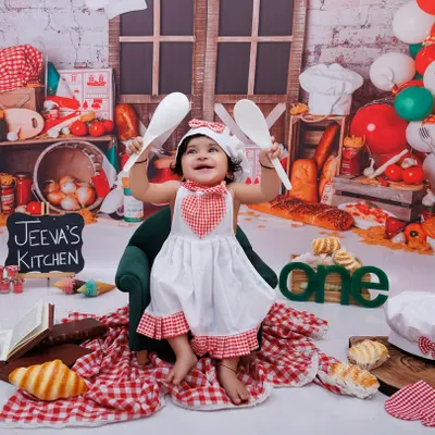Welcome to Jeeva's Kitchen! This little chef is ready to cook up some fun for her first birthday, holding up her spoons with excitement.
