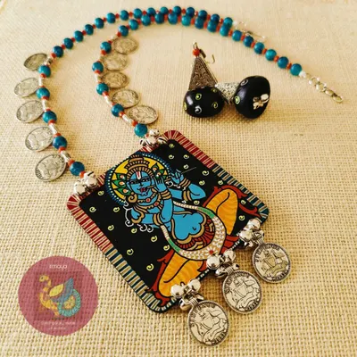 A hand-painted Sri Krishna Murari necklace, showing the lord in a classic pose. The MDF pendant is lacquer polished for a lasting finish and paired with wooden jhumkas, acrylic beads, and metal coin embellishments for a traditional feel.