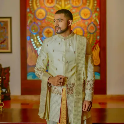 This is a custom-designed Indo-Western sherwani set. The off-white jacket features subtle embroidery on the sleeves and body, paired with a traditional dhoti and sash for a look that is both regal and contemporary.