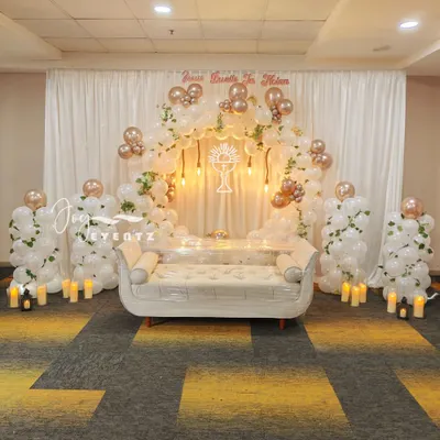 An elegant and expansive stage decoration for a First Holy Communion. This setup features a large balloon arch in white and rose gold, a comfortable white sofa, and a glowing chalice symbol at the center.