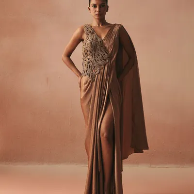 A modern interpretation of the sari, this draped gown in a muted gold tone features a thigh-high slit and a single flowing cape. The bodice is adorned with our signature cord work.