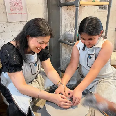 A mother and daughter sharing a smile at the potter's wheel. Our family workshops are designed to create these moments of pure joy and connection.