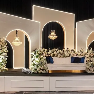 A modern and sophisticated stage design featuring geometric white panels with integrated lighting, grand chandeliers, and lush white floral arrangements for a luxurious feel.