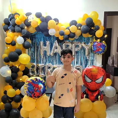 A happy birthday boy posing with his Spider-Man themed party decoration. This setup features a black and yellow balloon arch, a blue foil curtain, and character balloons of the friendly neighborhood superhero.