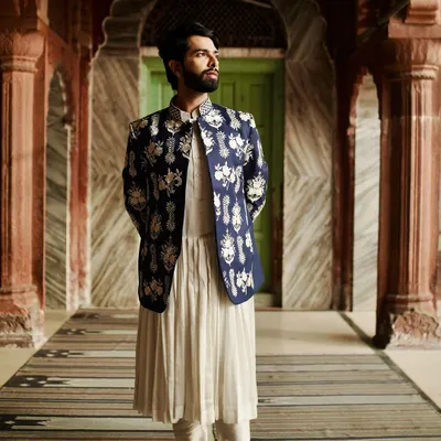 A full-length portrait showcasing a white pleated kurta layered with a navy blue Nehru jacket. The jacket is covered in intricate white floral embroidery, creating a look inspired by period dramas and royal aesthetics.
