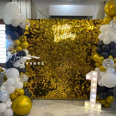 This is a classic and elegant first birthday setup featuring a gold shimmer wall. The backdrop is complemented by a 'Happy Birthday' neon sign and a beautiful balloon garland in black, white, and gold.