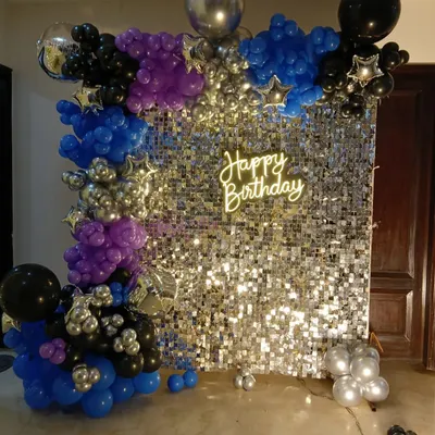 A vibrant party setup with a silver sequin wall. The backdrop is framed by a balloon garland in blue, purple, and black, and features a 'Happy Birthday' neon sign for a disco-ready vibe.