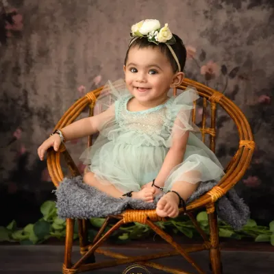 A perfect sitter session portrait. This happy baby girl is smiling brightly while sitting in a small wicker chair against a classic, painterly backdrop.