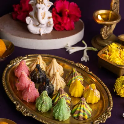 A vibrant platter of assorted modaks for Ganesh Chaturthi, styled with a Ganesha idol and hibiscus flowers for a devotional feel.