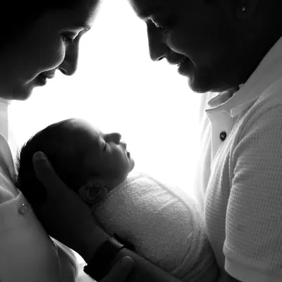 An intimate black and white silhouette of both parents gazing down at their newborn baby held lovingly in their hands.