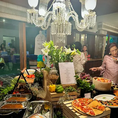 An elegant indoor setup for a house party. The buffet table is lavishly decorated with a crystal chandelier overhead, fresh white lilies, and our signature wooden crates, creating a luxurious feel while guests enjoy fresh pizza and other dishes.