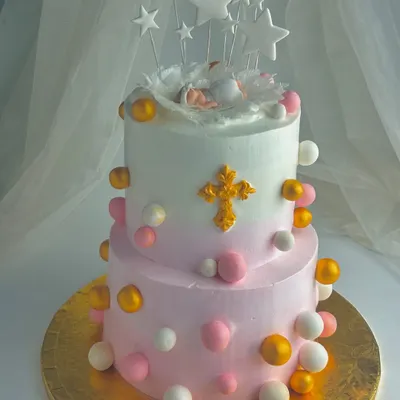 A beautiful two-tier cake for a child's dedication ceremony. The design features a sleeping baby in an angel's nest, a golden cross, and soft pink and white spheres, symbolizing a precious gift from above.