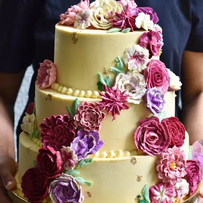 A stunning three-tiered wedding cake featuring a cascade of hand-piped buttercream flowers in shades of purple and pink. We specialize in creating these showstoppers that taste even better than they look.