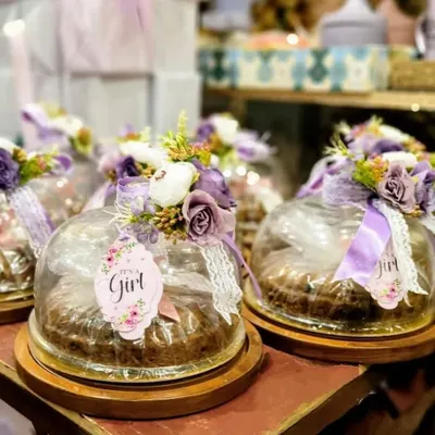 A close-up of our popular baby girl announcement favors. Each cake is presented under a protective dome and beautifully decorated with a posy of purple and white flowers, a lace ribbon, and a custom "It's a Girl" tag, making for a truly special keepsake gift.