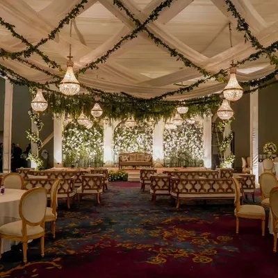 A breathtaking reception hall designed as an enchanted forest. The ceiling is draped with fabric and hanging greenery, with crystal chandeliers providing a warm, magical glow over the elegant seating arrangement.