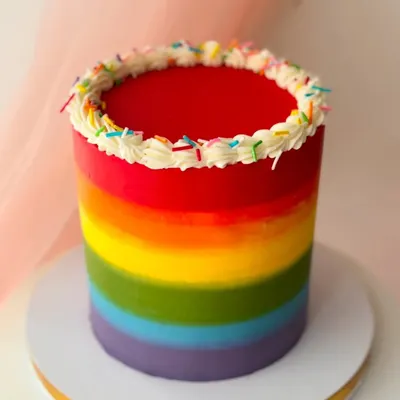 A vibrant rainbow cake with a bright red top. The smooth gradient is created with buttercream, and the top is finished with a simple piped border and sprinkles.