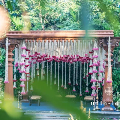 A breathtaking view of our summer wedding mandap, framed by lush greenery. The structure, made of intricately carved South Indian pillars, is adorned with cascading garlands of tuberose and pink oleander, creating a truly romantic and sacred space.