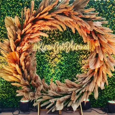 A rustic and chic photo booth for an engagement or reception. The backdrop features a large wreath made of pampas grass against a green wall, with a custom neon hashtag for the couple.