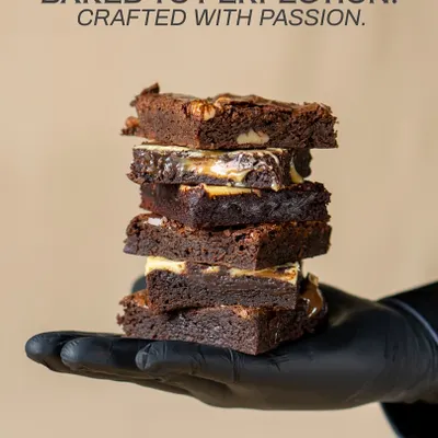 Baked to perfection, crafted with passion. A stack of my assorted brownies held in a gloved hand, showcasing the different textures and fillings I create.