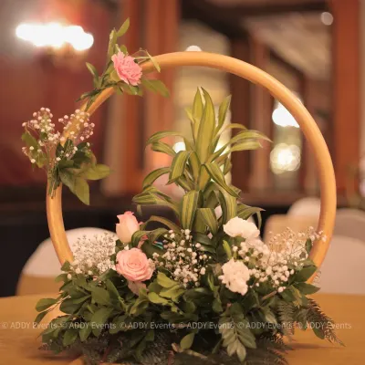 This modern centerpiece features a golden circular frame adorned with soft pink roses, white carnations, and baby's breath. It is a perfect example of how we blend contemporary design with classic floral beauty for table decor.