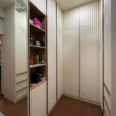 A walk-in closet space featuring floor-to-ceiling wardrobes with a unique curved fluting design on the doors. The design incorporates both closed storage and open shelving for display.