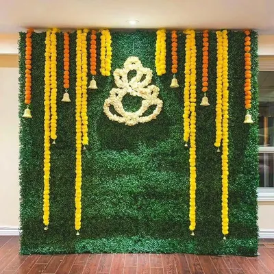 A beautiful and traditional backdrop for a religious ceremony, featuring a green wall decorated with marigold garlands and a central Ganesha motif made of white flowers.