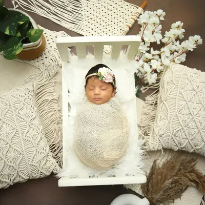 A beautifully styled shot featuring a newborn in a miniature white crib. The boho theme with macrame pillows and pampas grass creates a soft, neutral, and trendy look.