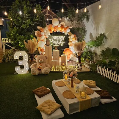 A cozy and chic teddy bear picnic theme for a 3rd birthday. The outdoor setup includes low seating with cushions, a neutral-toned balloon arch, and a light-up number.