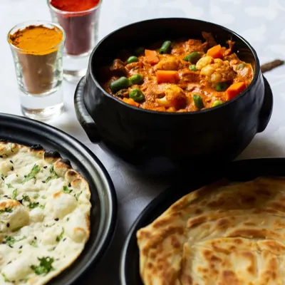 A wholesome North Indian meal featuring a rich vegetable curry, served with soft naan and flaky paratha. The small glasses contain the essential spices that give our curries their authentic flavor.