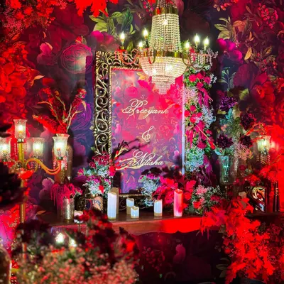 The welcome signage for a grand Sangeet, framed by an ornate gold mirror and surrounded by a sea of red florals and candles. A crystal chandelier adds a touch of regal elegance to the entrance vignette.