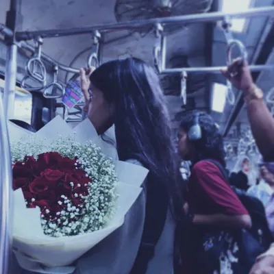 A quiet moment amidst the chaos of a Mumbai local train. A young woman holds a bouquet of red roses, lost in thought, showing how personal stories unfold even in the most public spaces.