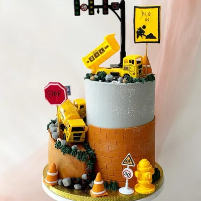 A two-tiered construction-themed cake with incredible detail. It features a winding road with toy trucks, traffic signs, and a "Dig In" sign, all on a cake designed to look like concrete and brick.