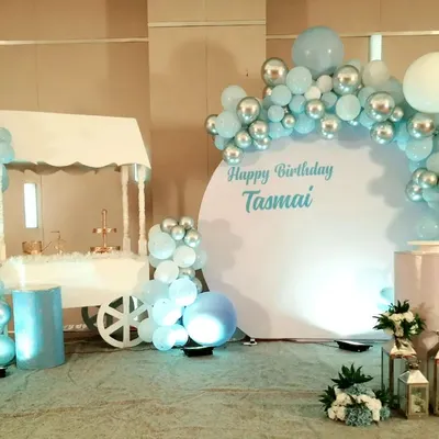 This modern and chic birthday backdrop features a clean white circular panel, perfect for custom text like 'Happy Birthday Tasmai'. The design is complemented by a stylish balloon half-arch in pastel blue, white, and silver chrome for an elegant look.