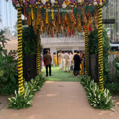 A festive entrance for a Mehndi ceremony. The arch is decorated with yellow and white flowers in a spiral pattern, with traditional marigold tassels hanging from the top to welcome guests.