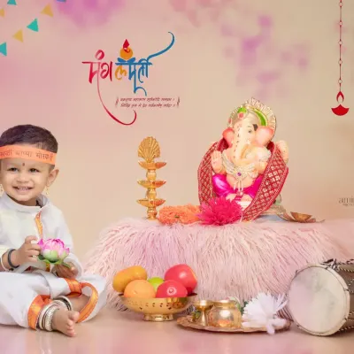 A smiling baby boy dressed in a white kurta with a "Ganpati Bappa Morya" headband. The setup includes a Ganpati idol, a dholak, and a bowl of fruits, capturing the complete festive vibe.