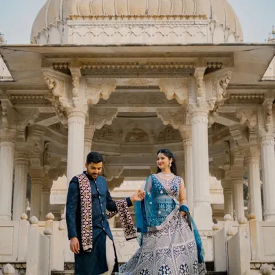 A classic shot of a couple descending the steps of a marble cenotaph at Gaitor Ki Chhatriyan. The groom gently leads the bride, showcasing the intricate details of their traditional blue outfits against the white marble.