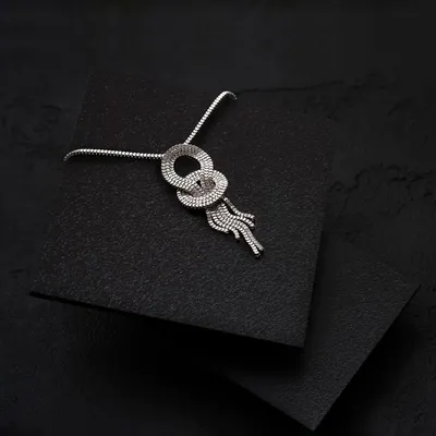 An elegant silver necklace with a knotted design, photographed on a textured black surface. The minimalist setup and focused lighting draw all attention to the product's unique form and craftsmanship.