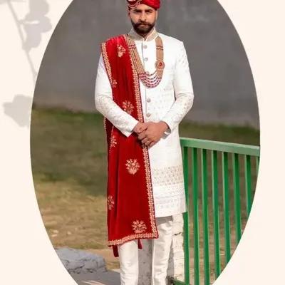 A groom looking sharp in a classic ivory sherwani paired with a rich red velvet stole. The intricate embroidery and matching red pagri create a regal and coordinated look.