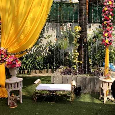 A bright and cheerful photo booth designed for a Haldi ceremony. The setup includes a comfortable bench set against a backdrop of vibrant yellow drapes, floral arrangements, and hanging lights.