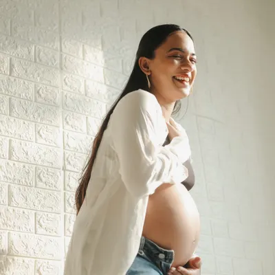 A joyful and candid lifestyle portrait. The mom-to-be is dressed in jeans and a simple white shirt, laughing naturally in the warm sunlight of our studio.