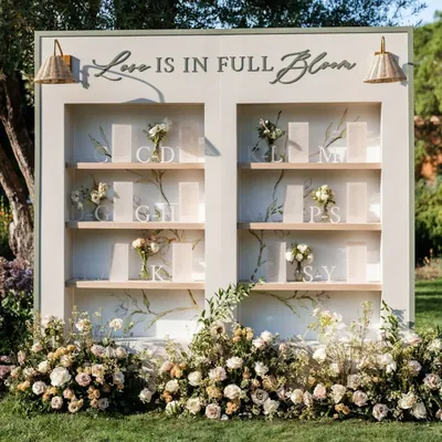 "Love is in Full Bloom." This custom seating chart display was designed for a garden wedding, with guest lists presented on elegant shelves adorned with fresh, delicate flowers.