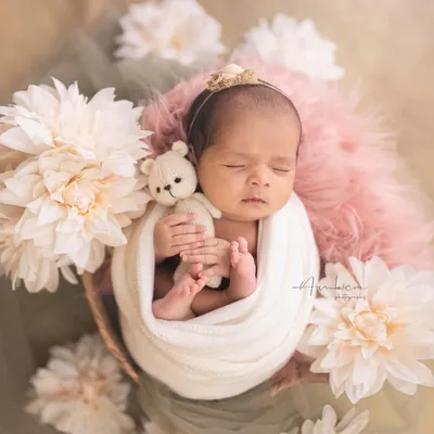 This is the essence of a classic newborn portrait. The baby is swaddled snugly, holding a tiny bear, and surrounded by soft flowers, creating a dreamy and peaceful image.