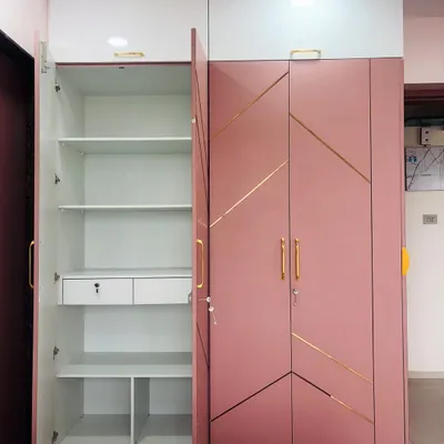 This pink and white hinged wardrobe showcases a clean, modern aesthetic with gold-accented geometric lines. The open door reveals the highly functional interior, complete with adjustable shelves, drawers, and dedicated compartments for organized storage.